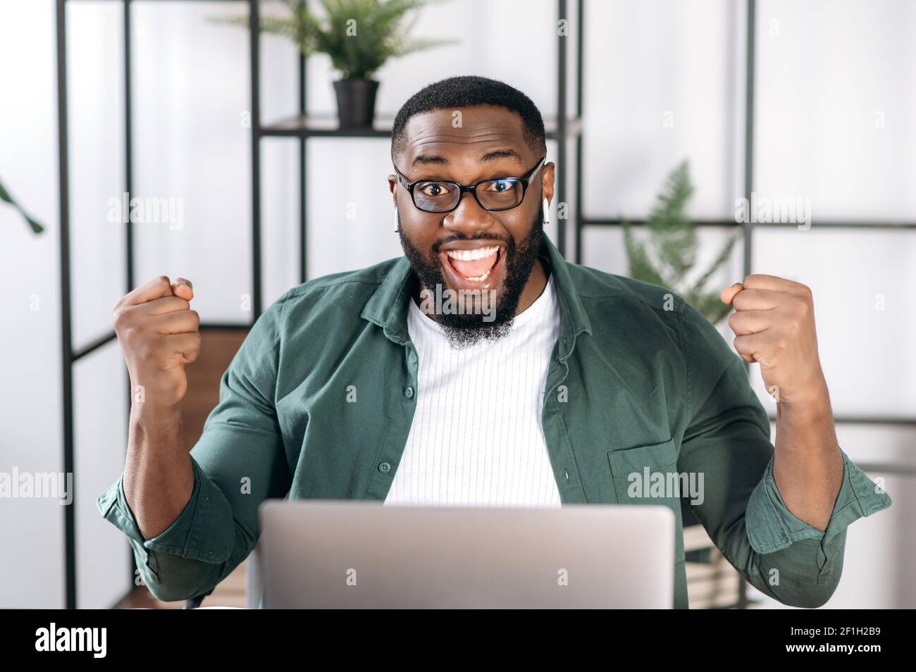 Esultante freelancer africano-americano vestito con abiti eleganti, seduto alla sua scrivania, gioendo nel successo, vittoria o buon profitto, guardando la macchina fotografica e gesturando con le mani Foto Stock