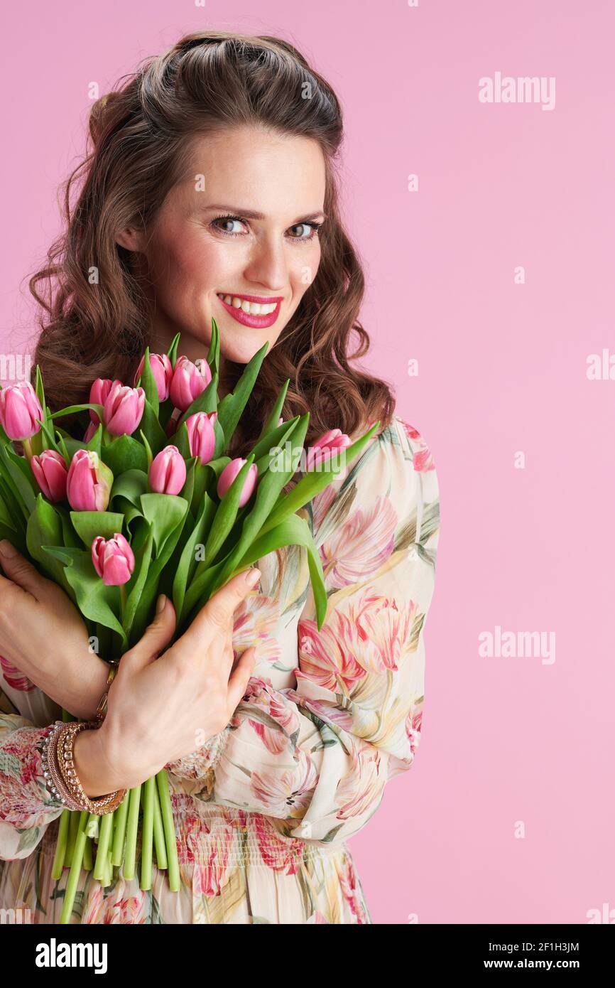 sorridente elegante donna di mezza età in abito floreale con bouquet di tulipani su sfondo rosa. Foto Stock