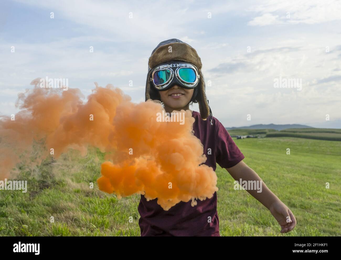 Immaginazione, ragazzo piccolo che indossa casco e oggetti d'oca in piedi su un campo verde con fumo colorato, fingendo di essere un pi Foto Stock