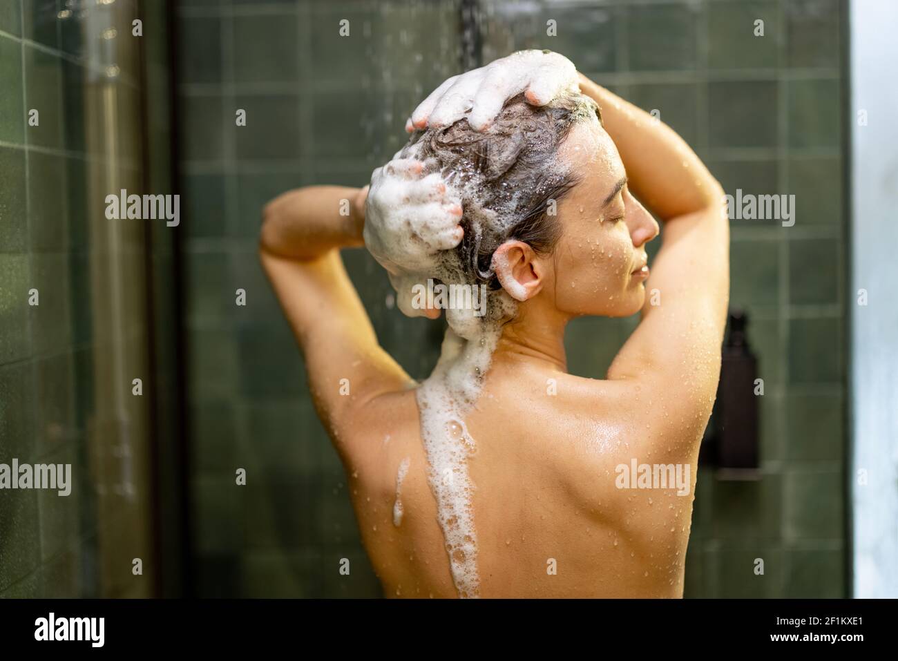 La donna che si ferma nella doccia applica lo shampoo con acqua  gocciolante. Fare la doccia e rilassarsi sotto l'acqua corrente calda.  Godetevi durante il bagno d'acqua calda con la schiuma nelle