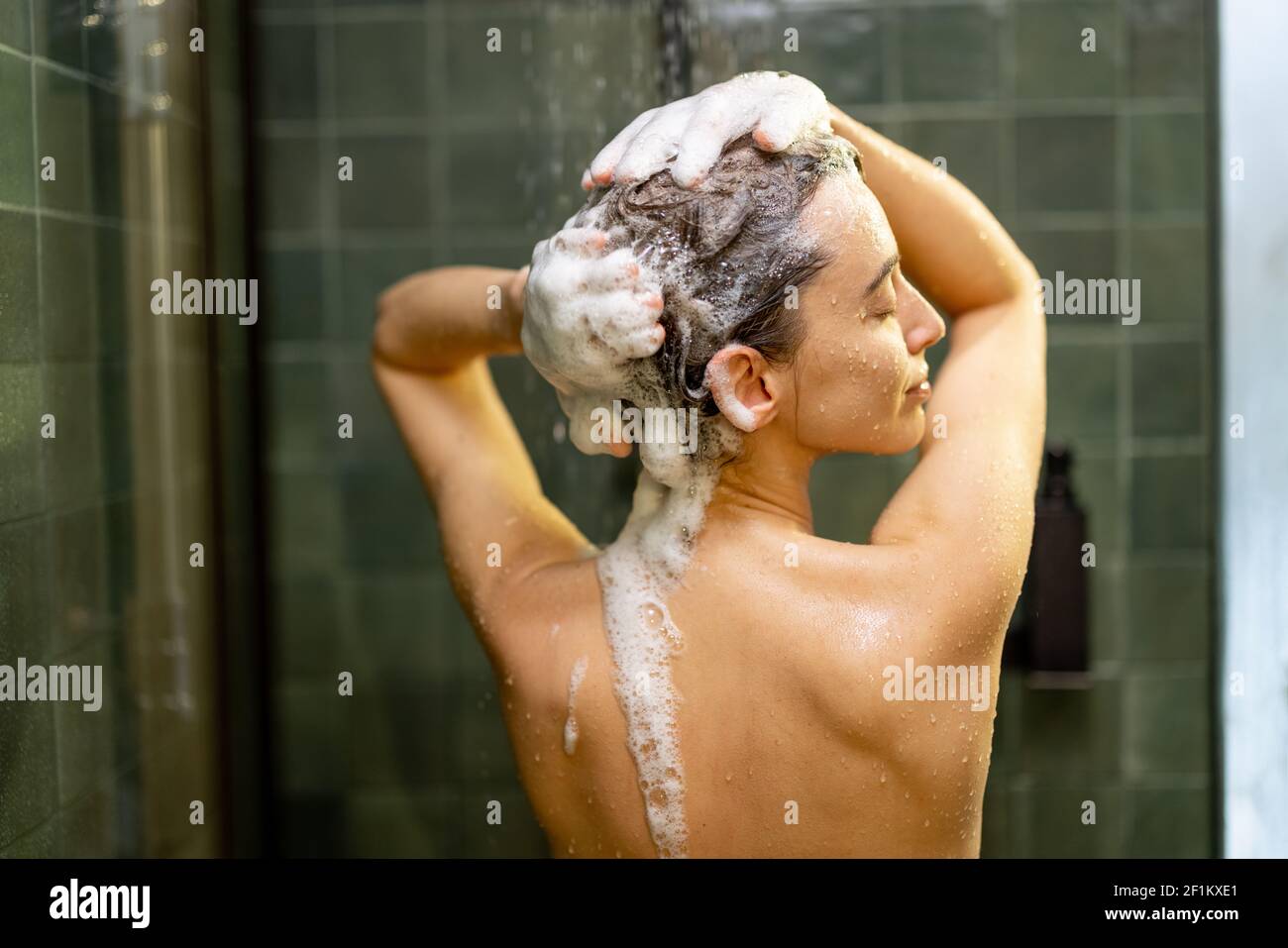 La donna che si ferma nella doccia applica lo shampoo con acqua gocciolante. Fare la doccia e rilassarsi sotto l'acqua corrente calda. Godetevi durante il bagno d'acqua calda con la schiuma nelle mani. Foto Stock