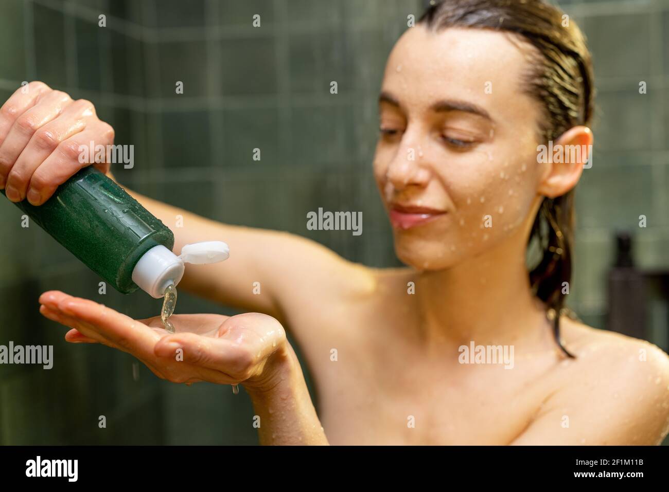 La donna prende lo shampoo dalla bottiglia verde, prendendo la doccia in bagno verde con acqua che gocciola sul viso. Primo piano. Capelli bagnati pettinati indietro. Foto Stock