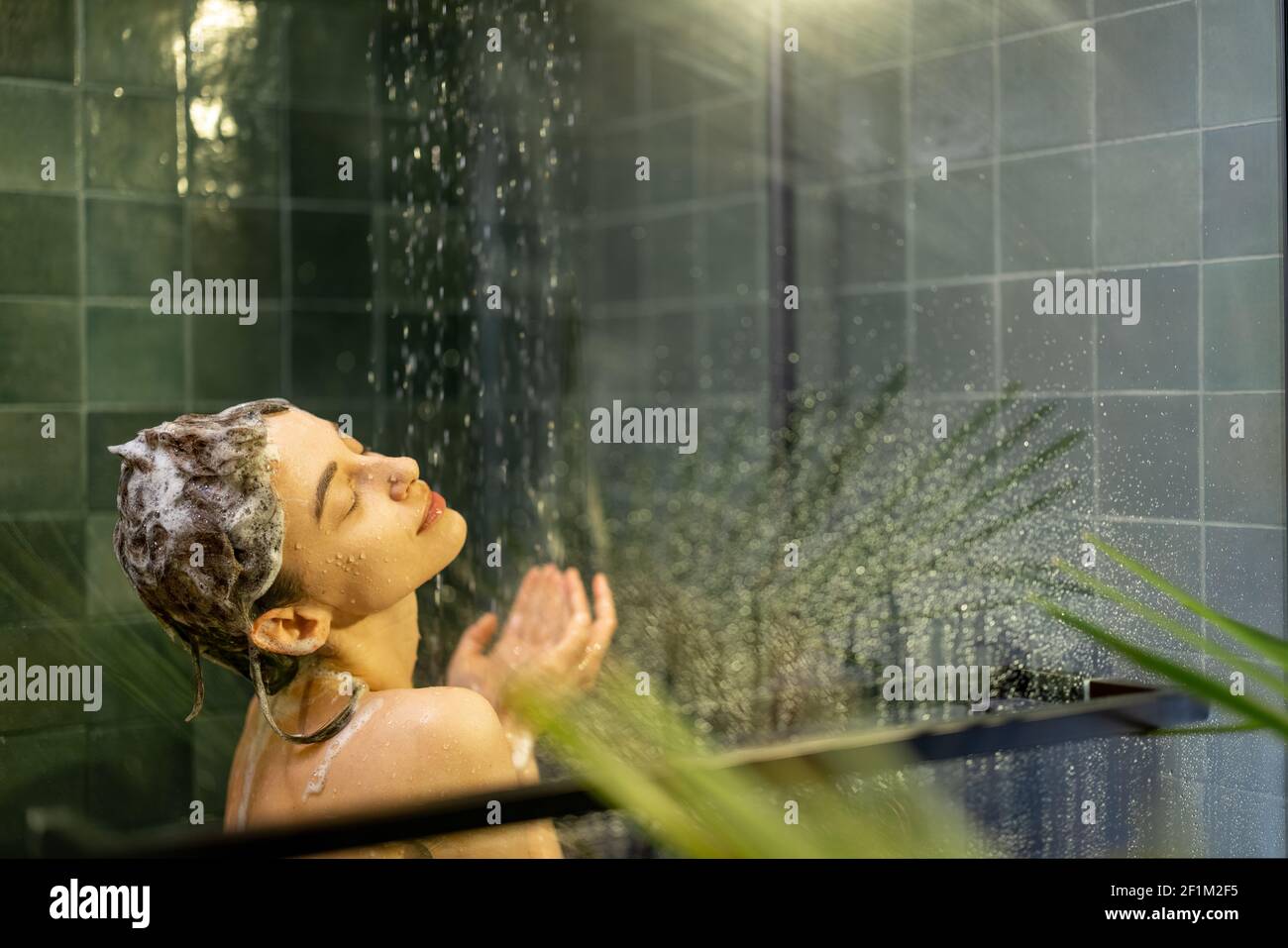 La donna che si ferma nella doccia applica lo shampoo con acqua che gocciola accanto alla parete di vetro. Prendere la doccia e rilassarsi sotto l'acqua corrente calda nel verde fresco bagno design. Spazio di copia. Foto Stock