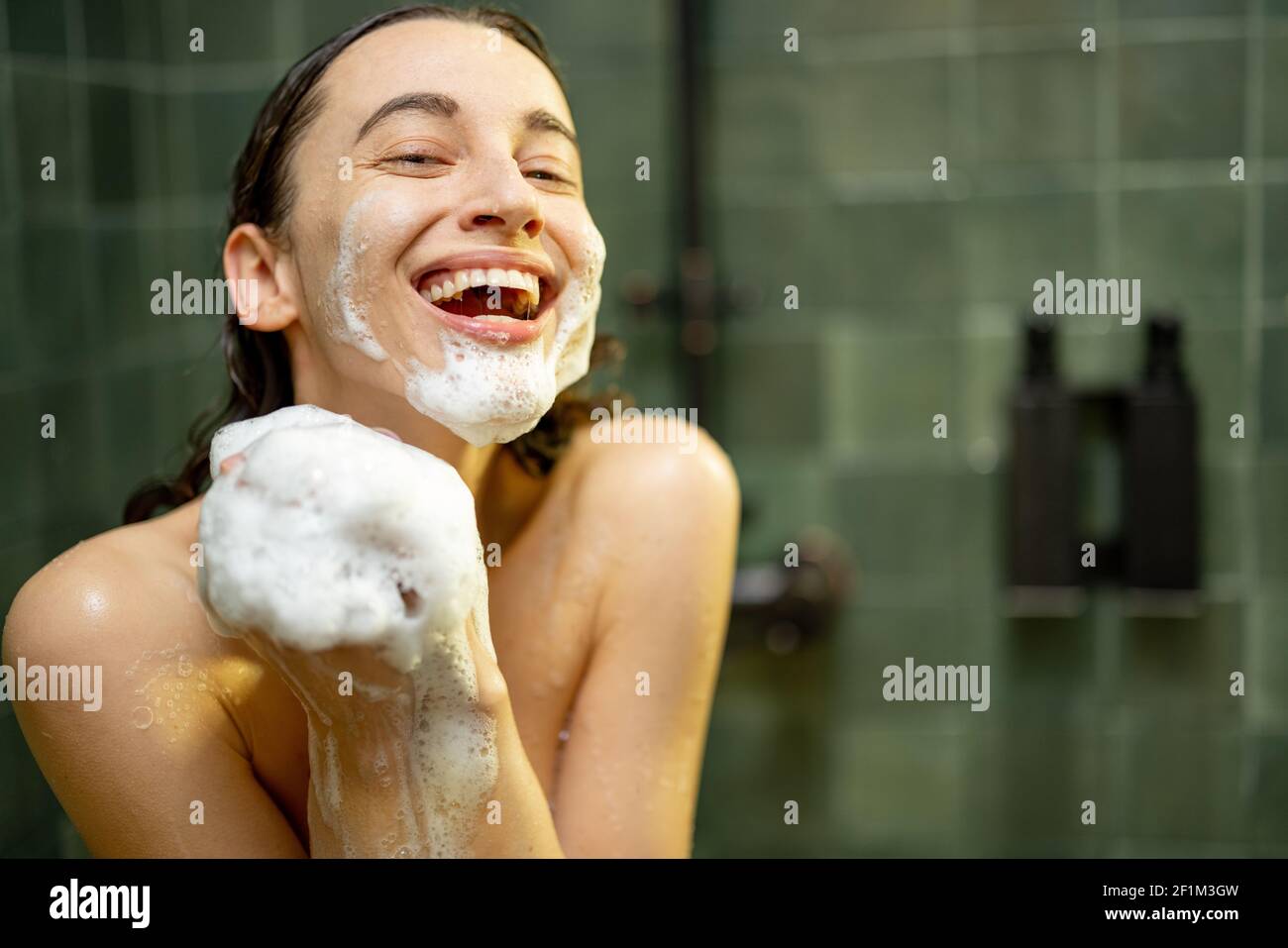 Donna sorridente che prende la doccia e che sorvola il corpo con gel doccia in bagno verde. Foto di routine quotidiana. Guardando la fotocamera. Foto Stock
