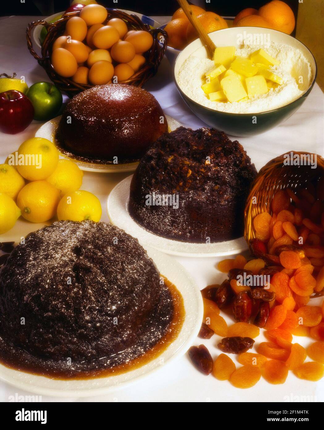 3 budini cotti su piatti bianchi con ingredienti uova, burro, farina, limoni, mele, albicocche secche, datteri su fondo bianco recipiente di miscelazione Foto Stock