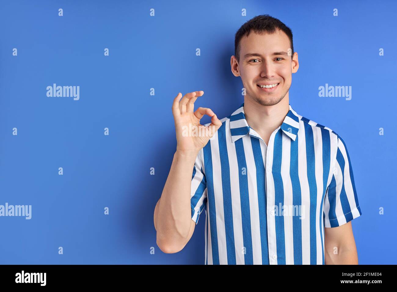 Uomo allegro che mostra un gesto e un sorriso OK, si sente felice, di buon umore. Giovane ragazzo di successo in camicia a righe blu che posa isolato su sfondo blu, Foto Stock