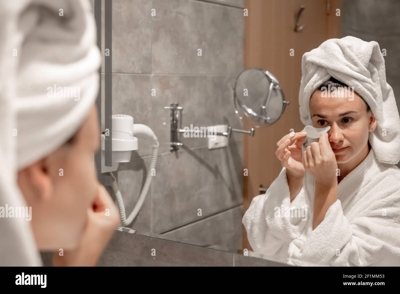 Una ragazza un accappatoio e con un asciugamano sulla sua testa bastoni macchie sotto i suoi occhi nel bagno di fronte allo specchio. Foto Stock