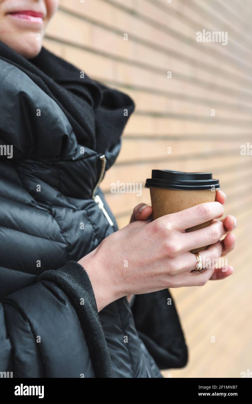 vista verticale della tazza di caffè usa e getta trattenuta dalle mani di una giovane caucasica irriconoscibile. Sta vestendo il cappotto nero elegante e in piedi vicino ad una parete di mattone Foto Stock