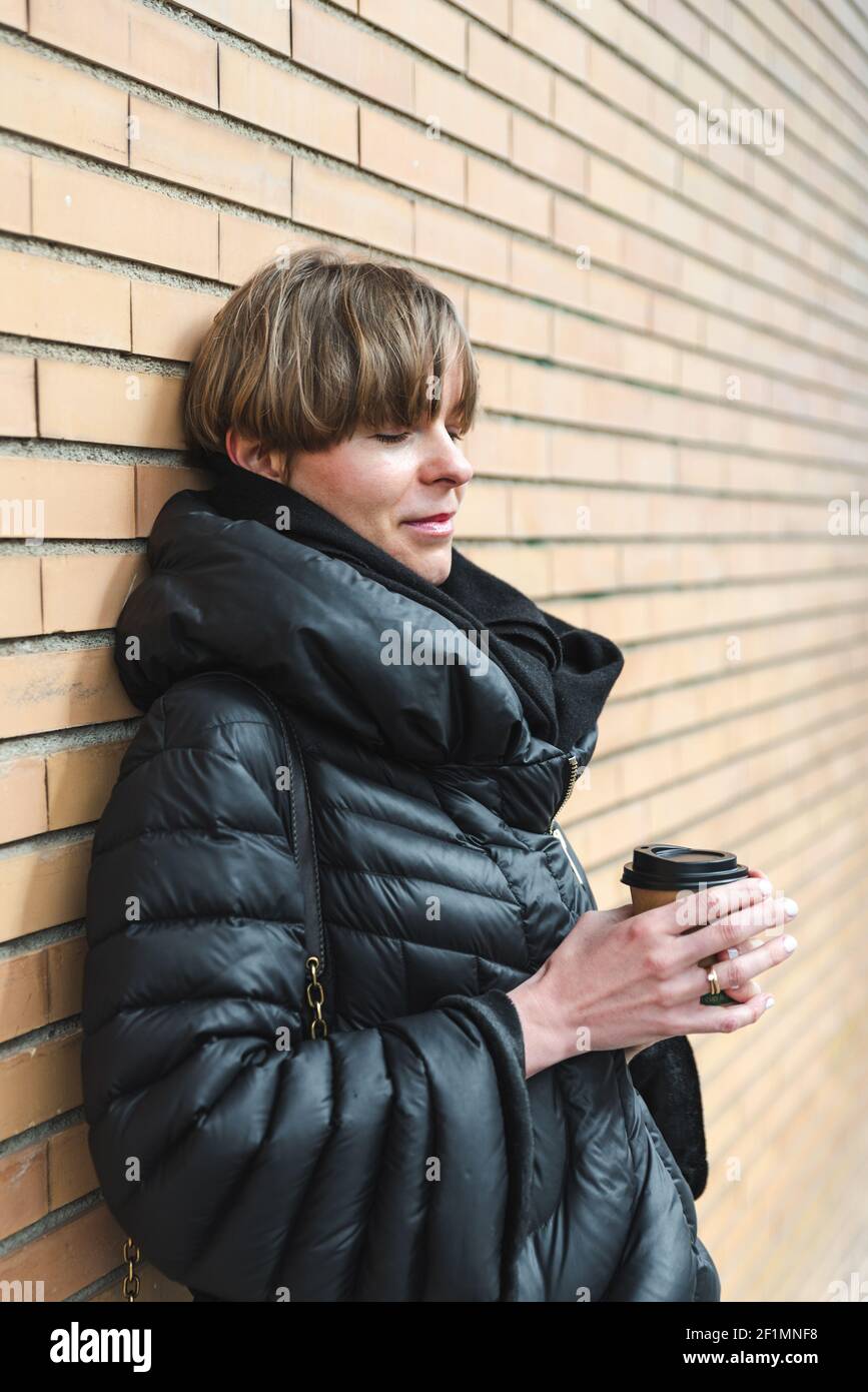 bionda giovane bella donna vestendo cappotto nero e tenendo una tazza di carta di caffè take away vicino ad una parete fatta di mattoni arancioni. Spazio di copia a destra Foto Stock