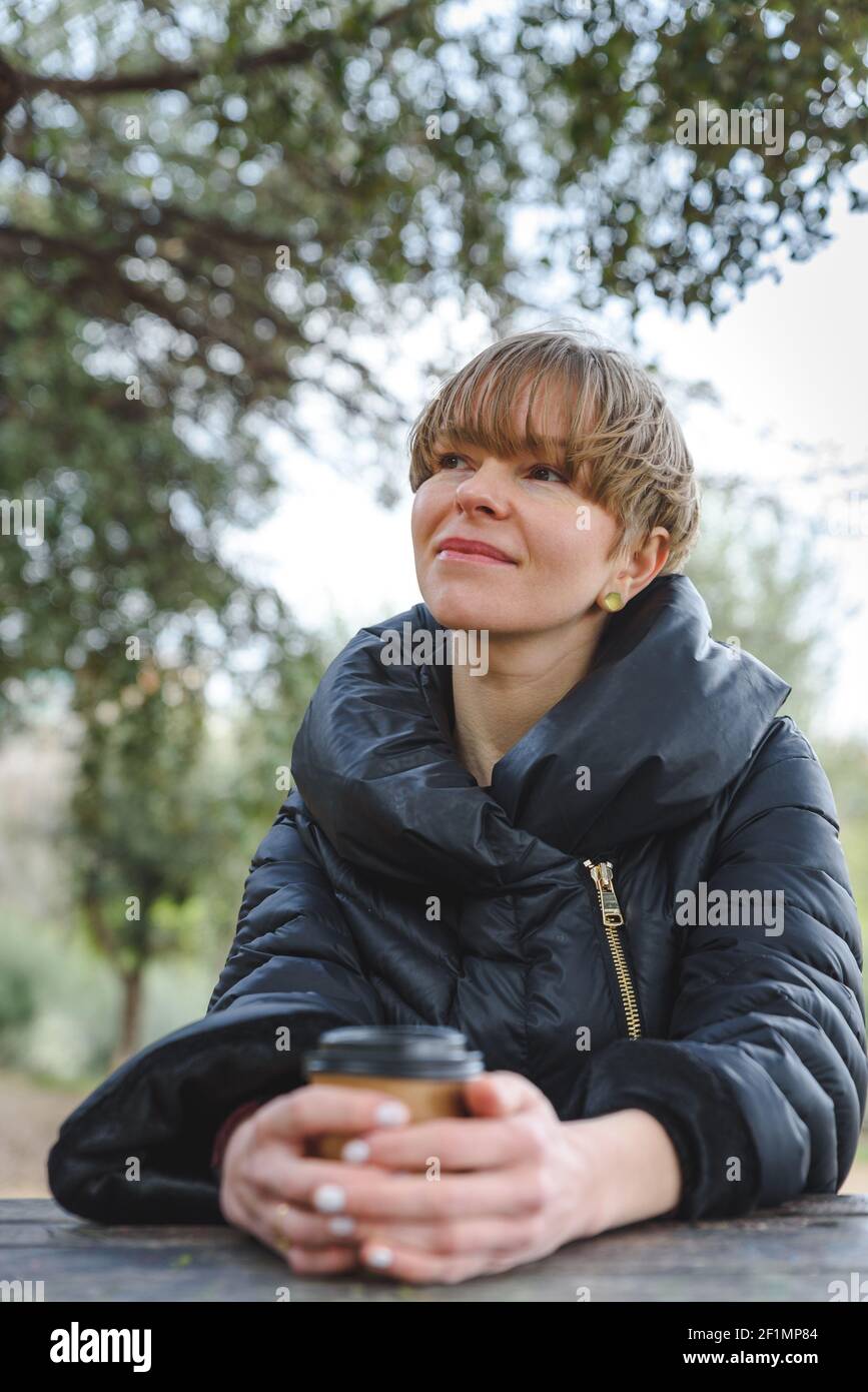 bella giovane bionda seduta vicino ad un tavolo di legno in un parco verde. Indossa un cappotto nero alla moda e ha una tazza di caffè di carta usa e getta sulle sue mani. Ha un aspetto ed un'espressione ponderati. Foto Stock