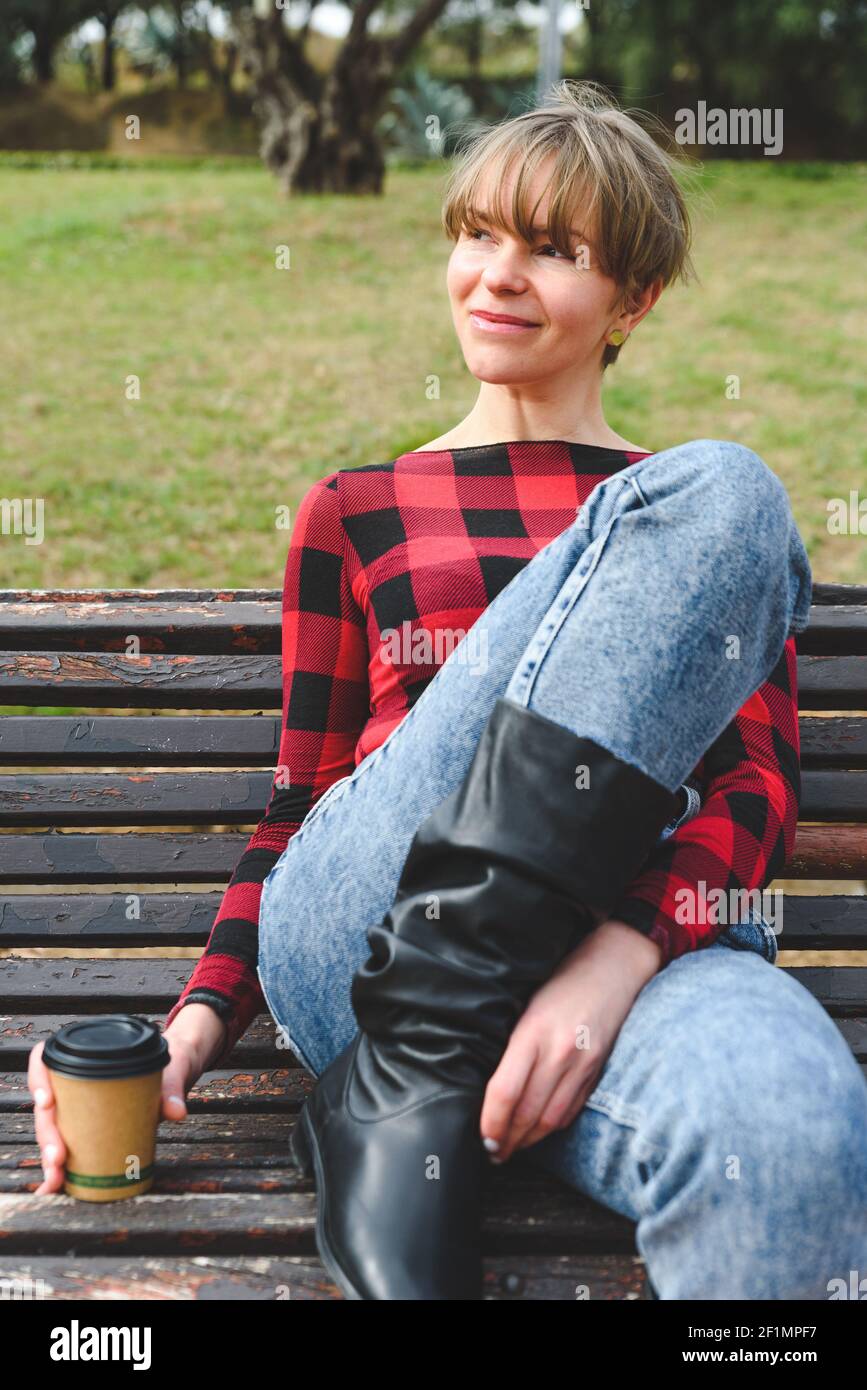 ritratto verticale di un modello biondo seduto su una panchina in un parco urbano verde e tenendo una tazza usa e getta con caffè. Sta vestendo i vestiti casual, i jeans blu, la camicia quadrata rossa e nera del motivo e gli stivali lunghi eleganti Foto Stock
