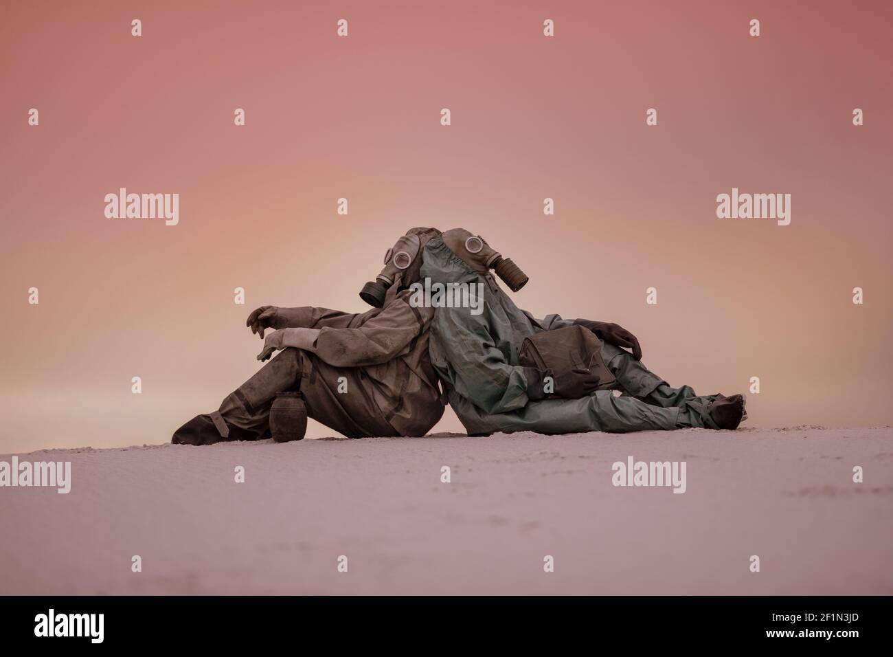 mondo post-apocalittico dopo una guerra nucleare. Due uomini in maschere a gas e tute protettive si siedono sulla sabbia con le spalle l'una all'altra. Foto Stock