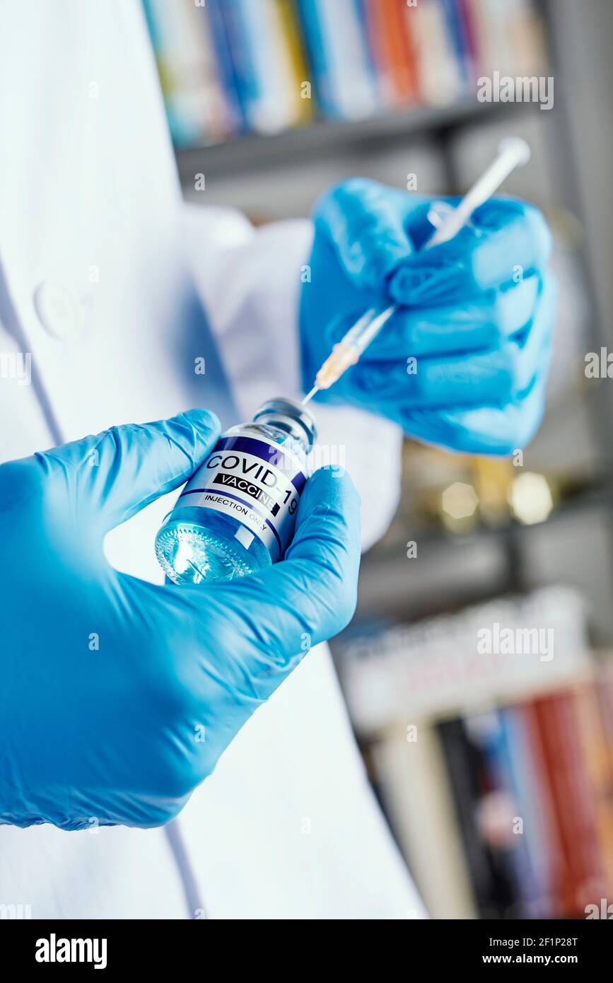 un operatore sanitario, indossando camice bianco e guanti chirurgici blu, riempie una siringa da un flaconcino simulato del vaccino covid-19, in una casa o in un nur Foto Stock