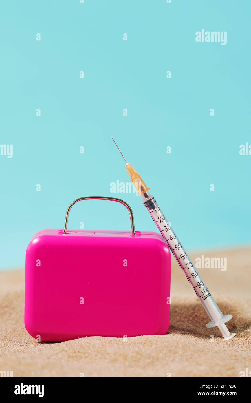una siringa e una valigia rosa sulla sabbia, su uno sfondo blu con uno spazio vuoto sulla parte superiore, raffigurante il settore del turismo medico Foto Stock
