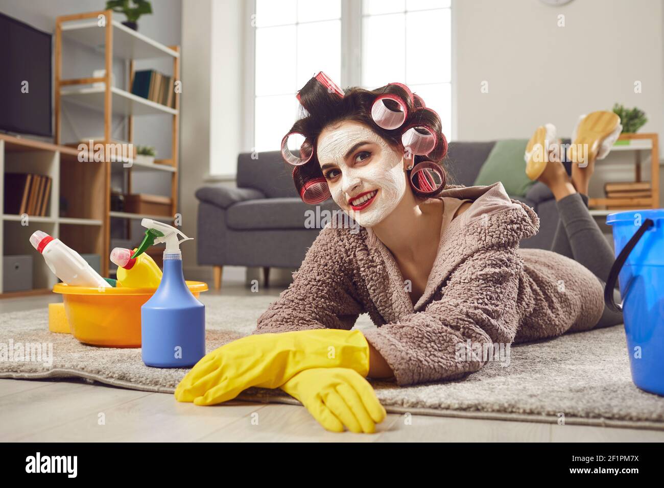 Donna felice in arricciacapelli, maschera facciale di bellezza e guanti di gomma che si rilassano dopo aver fatto tutto il lavoro domestico Foto Stock