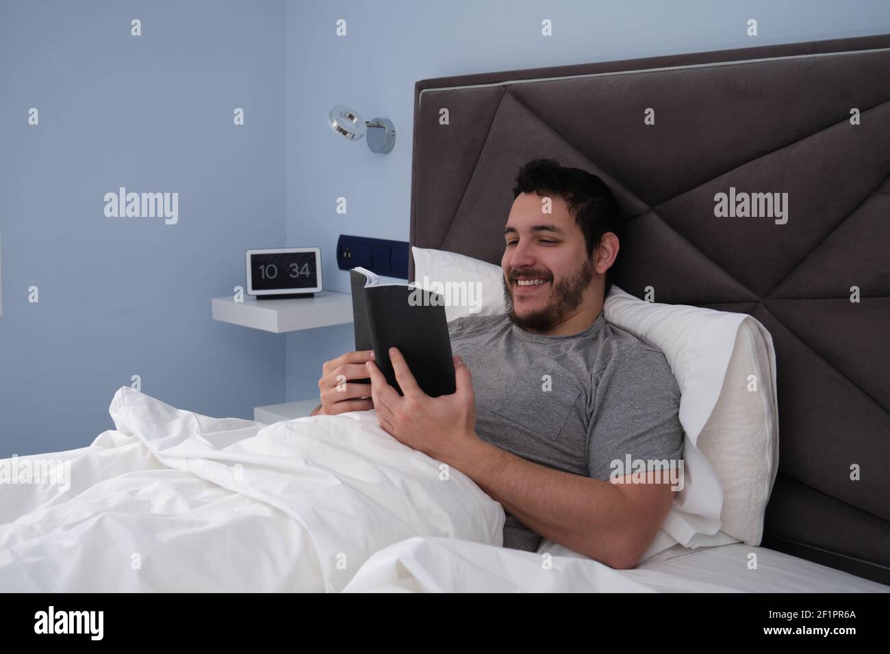 Giovane latino che legge un libro e sorride a letto a tarda mattinata. Orologio digitale in background. Foto Stock