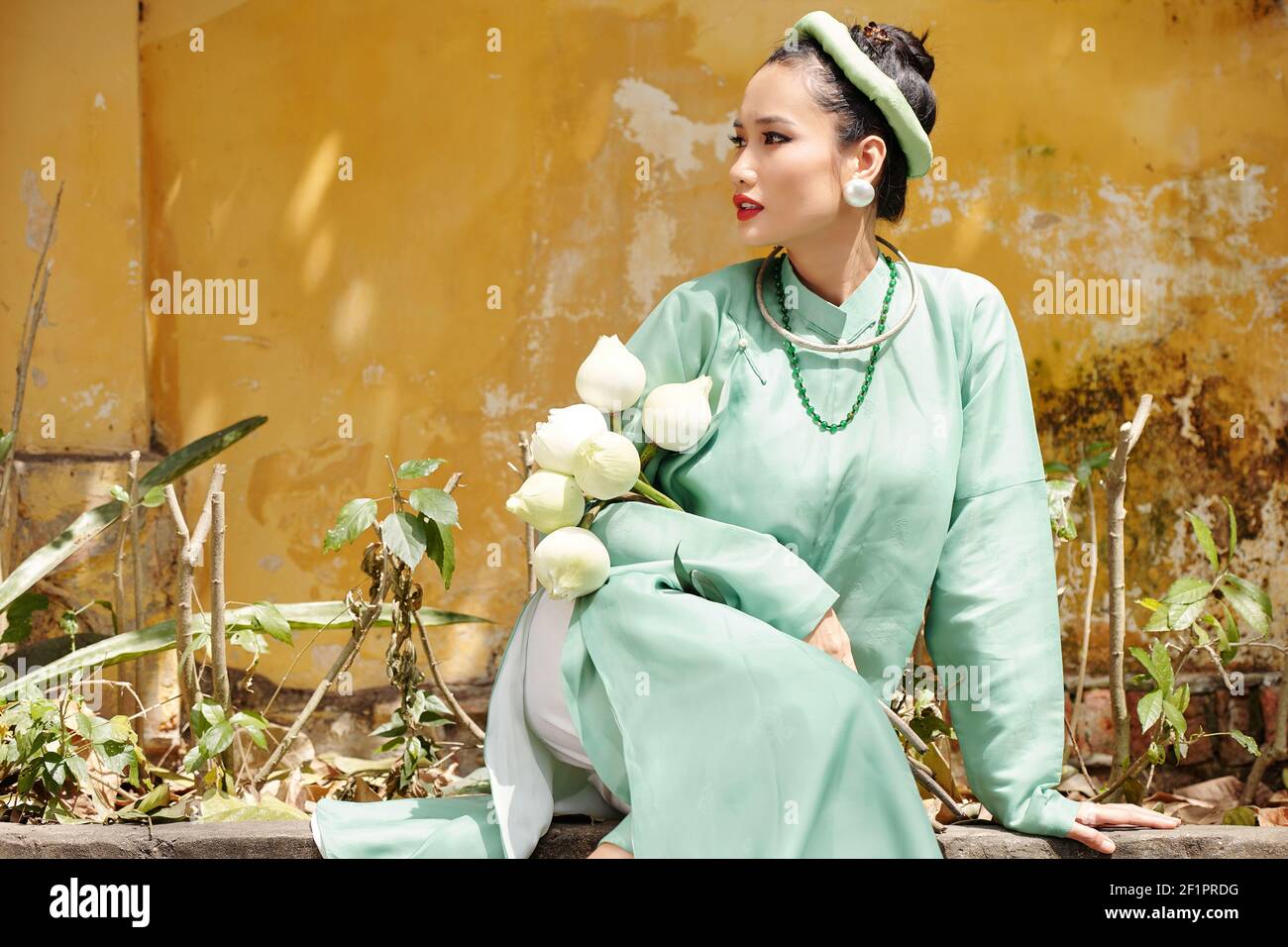 Attraente giovane donna vietnamita in verde menta ao dai vestito e cappelli tradizionali seduti all'aperto con fiori di loto in mani Foto Stock