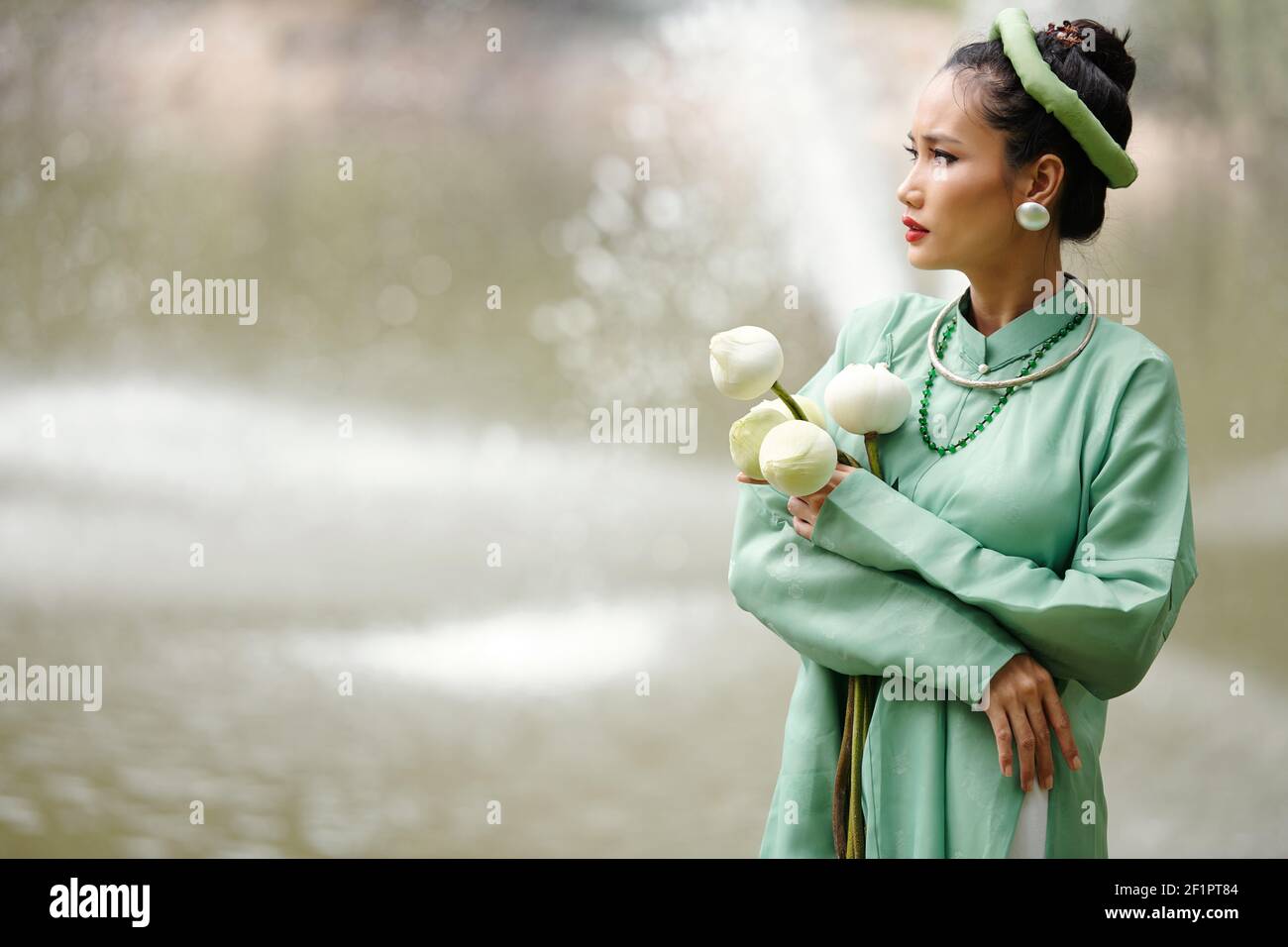 Preoccupata giovane donna vietnamita in costume tradizionale in piedi fiume con fiori di loto in mani e guardando l'acqua Foto Stock