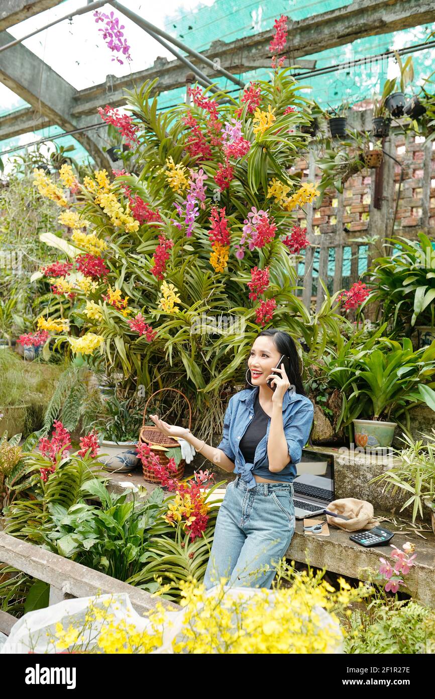 Felice giovane donna asiatica che lavora in serra e parla al telefono con il cliente o l'appaltatore Foto Stock