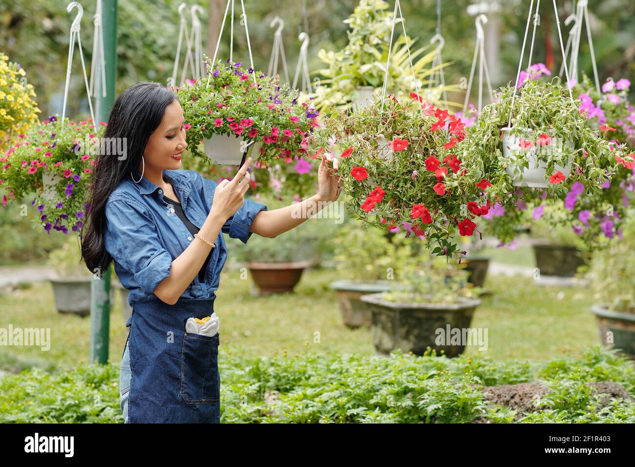 Bella sorridente giovane donna asiatica che lavora in vivaio di fiori e. fotografare fiori appesi per il suo negozio online Foto Stock