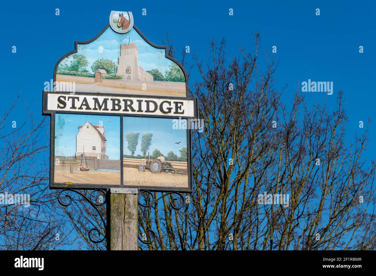 Segno del villaggio di Stambridge con immagini di caratteristiche locali, tra cui la chiesa di Santa Maria e tutti i Santi, trattore che coltivano un campo e un mulino Foto Stock