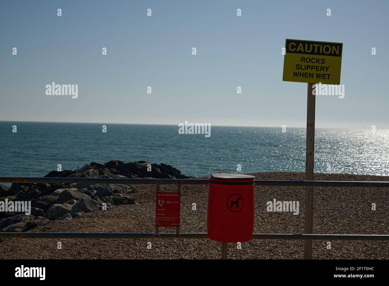 Un poo bin rosso cane, un segno sulle rocce scivolose e una recinzione di fronte a una spiaggia di ciottoli nel sud dell'Inghilterra. Cielo blu e orizzonte del mare. Foto Stock