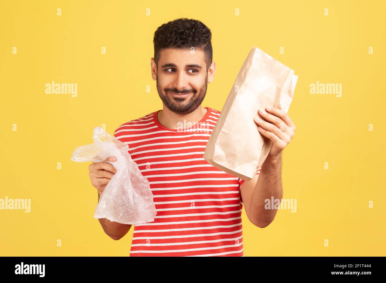 Felice uomo allegro con barba in t-shirt a righe rosse che tiene due borse in mano, guardando la borsa di carta con sorriso, rifiutando di usare la plastica. studi interni Foto Stock