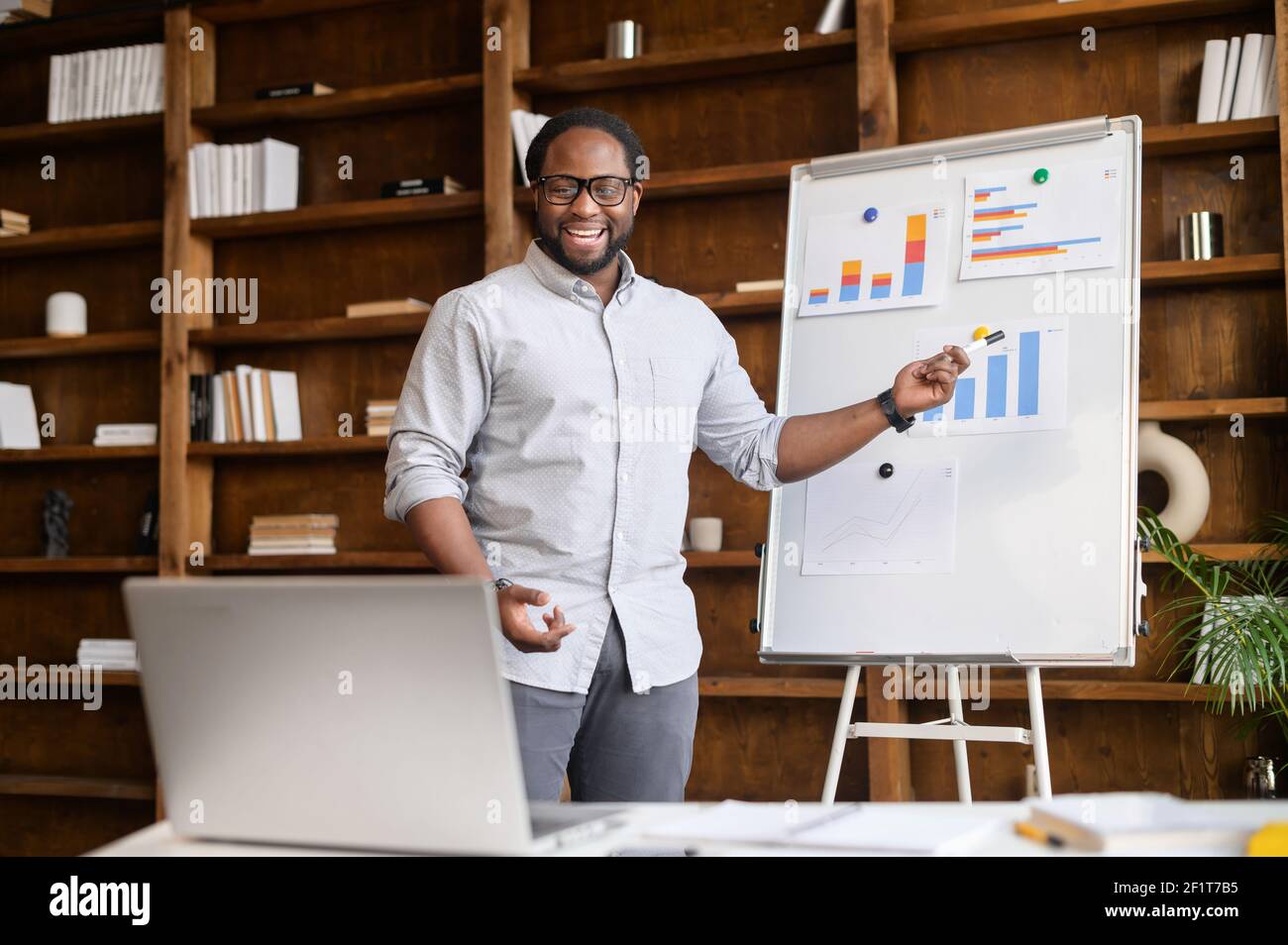 Un insegnante online afroamericano, un tutor, un business coach tiene un webinar, punta a lavagna bianca con grafici, spiega al pubblico online sul laptop, uomo biraciale fa una presentazione online Foto Stock