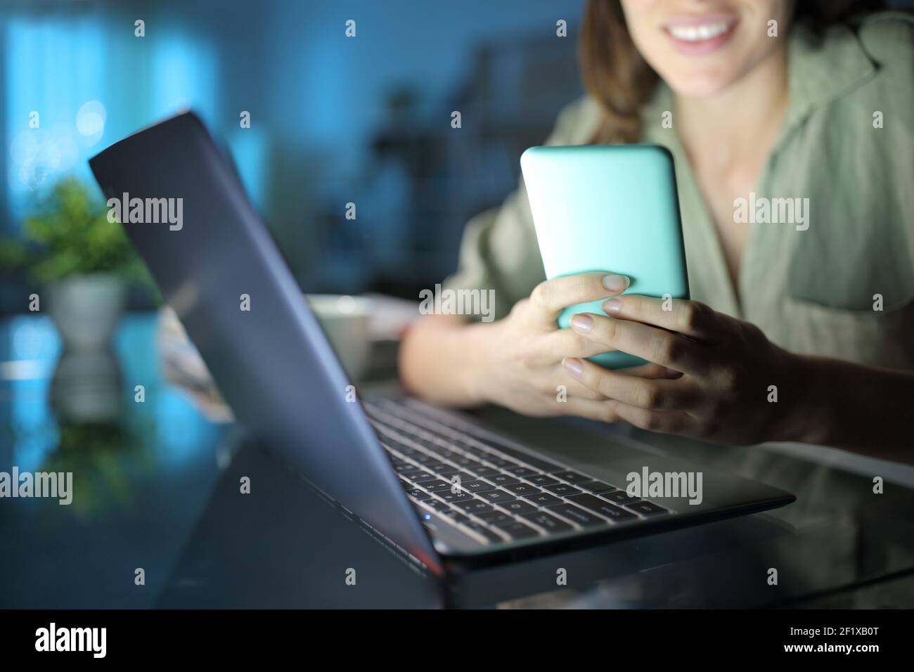 Primo piano di una donna felice che usa il SMS dello smartphone messaggio accanto a un computer portatile di notte a casa Foto Stock