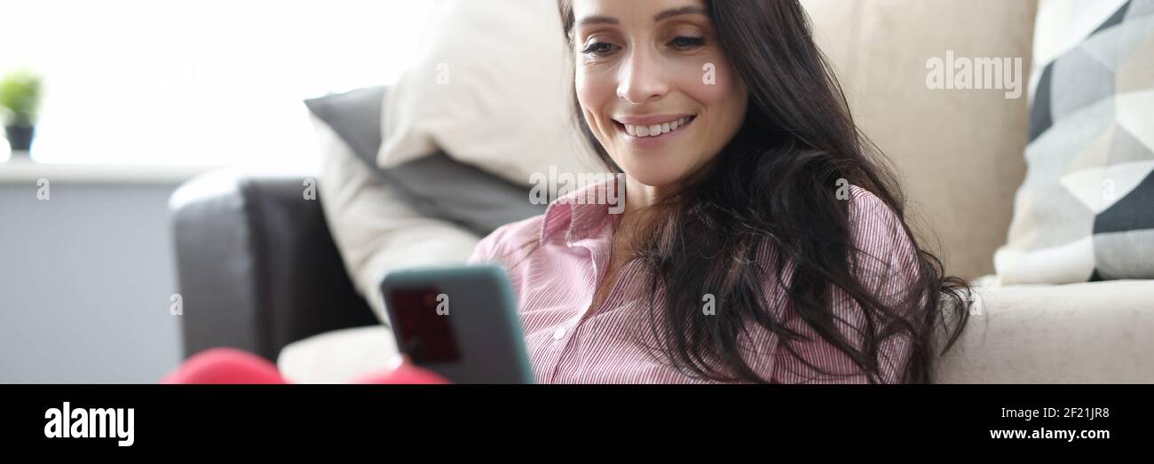 Donna si siede sul pavimento accanto al divano e guarda sorridente sullo schermo dello smartphone. Foto Stock
