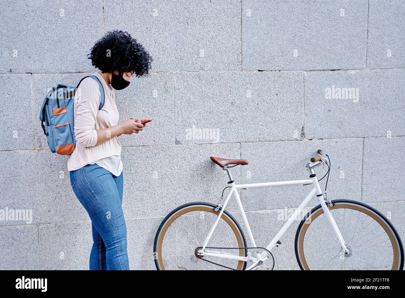 Donna afroamericana con uno smartphone in mano. Donna con maschera protettiva. Andare in bicicletta in città. Foto di alta qualità Foto Stock