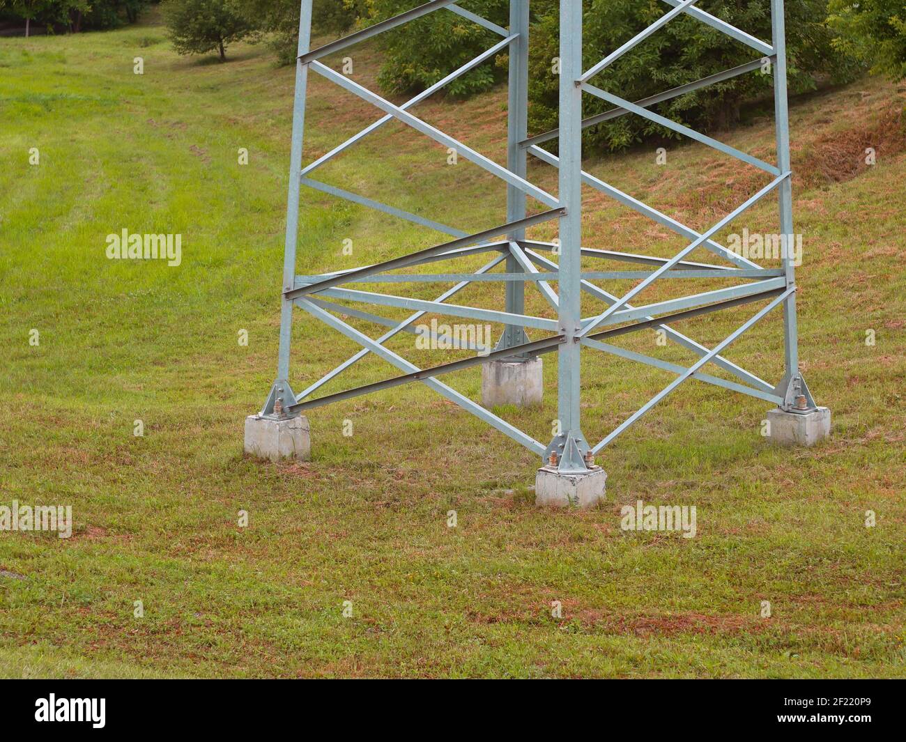 Parte inferiore di un grande supporto in acciaio elettrico di torre di trasmissione c.a. ad alta tensione su sfondo naturale, struttura metallica fissata ad una base in cemento Foto Stock