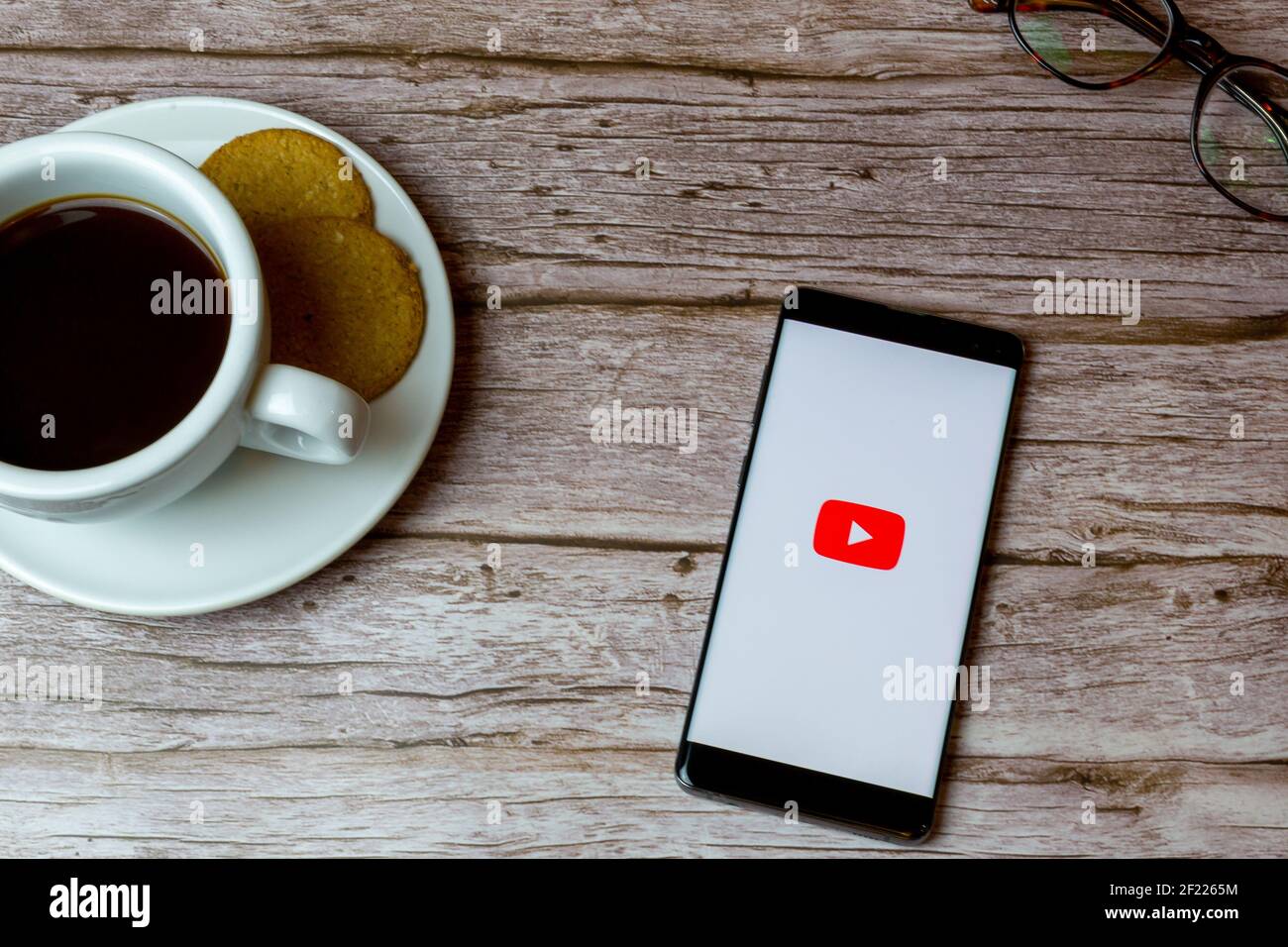 Un telefono cellulare o un telefono cellulare posato su un legno Tavolo con l'app YouTube che apre anche un caffè e. occhiali Foto Stock