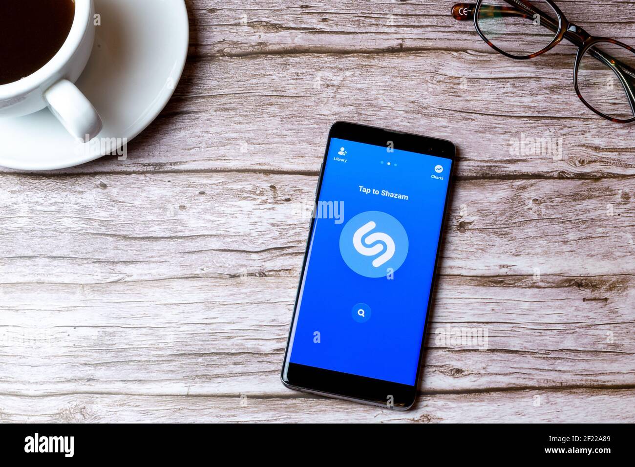 Un telefono cellulare o un telefono cellulare posato su un legno Tavolo con l'app Shazam aperta accanto a un caffè e bicchieri Foto Stock