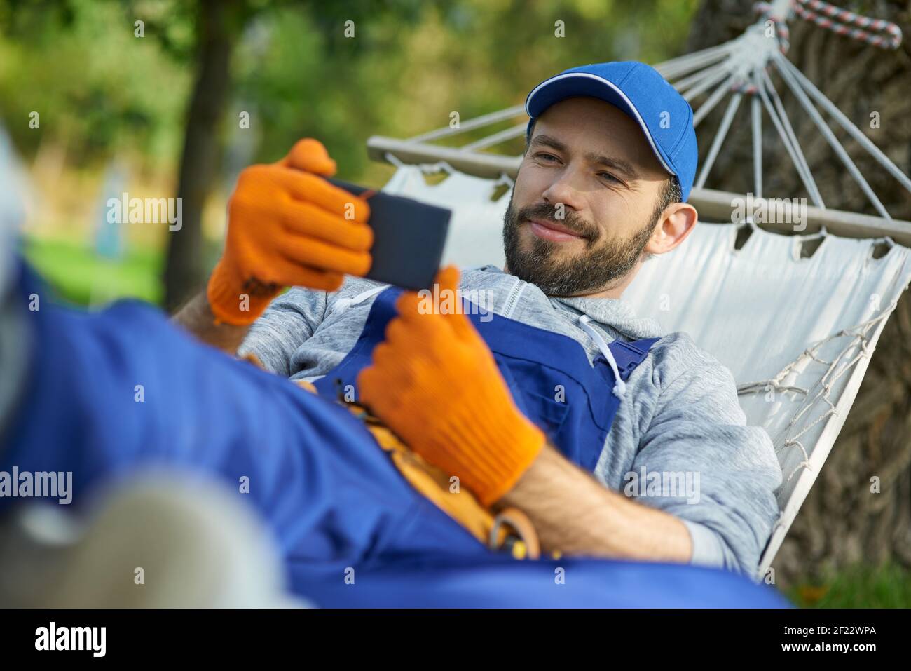 Felice giovane costruttore maschile indossare uniforme prendere una pausa, sdraiato in un amaca all'aperto e guardare qualcosa utilizzando smartphone in una giornata di sole. Costruzione, professione, concetto di riposo Foto Stock