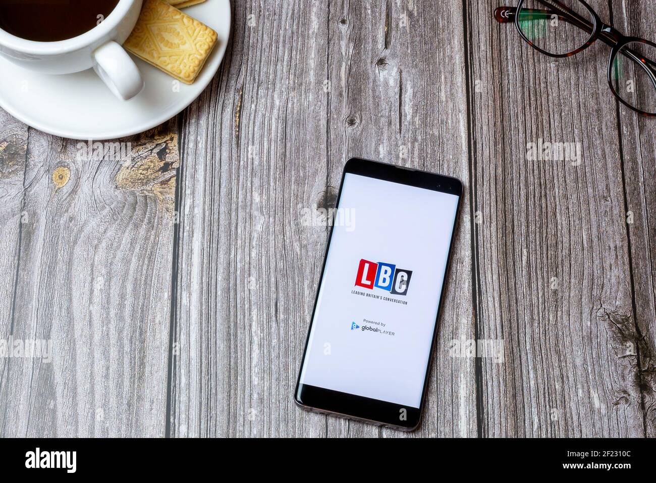 Un telefono cellulare o cellulare su un tavolo di legno Con l'app LBC radio aperta accanto a un caffè e bicchieri Foto Stock