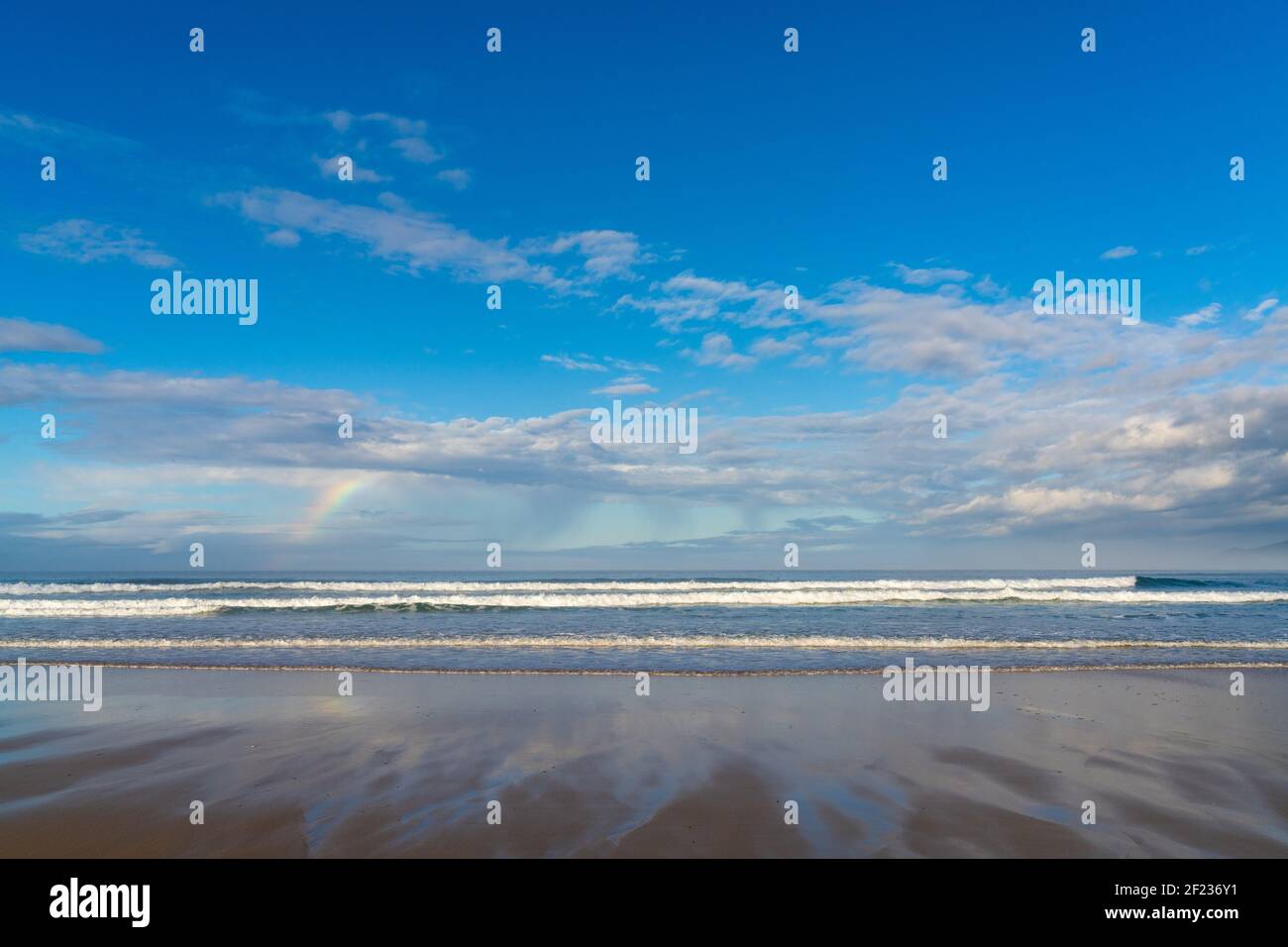 Una spiaggia di sabbia con grandi onde e un piccolo arcobaleno in un cielo espressivo Foto Stock