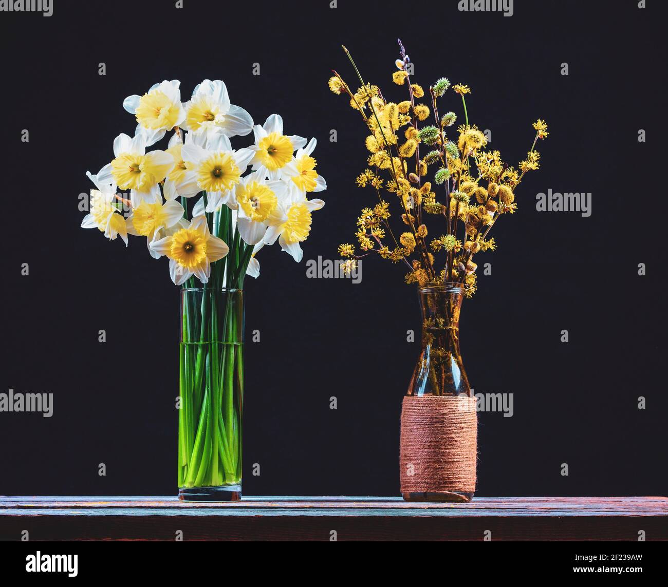 un bouquet di fiori di narcisi e un bouquet di fiori salice e dogwood rami con fiori gialli in vasi di vetro su un tavolo di legno su un b nero Foto Stock