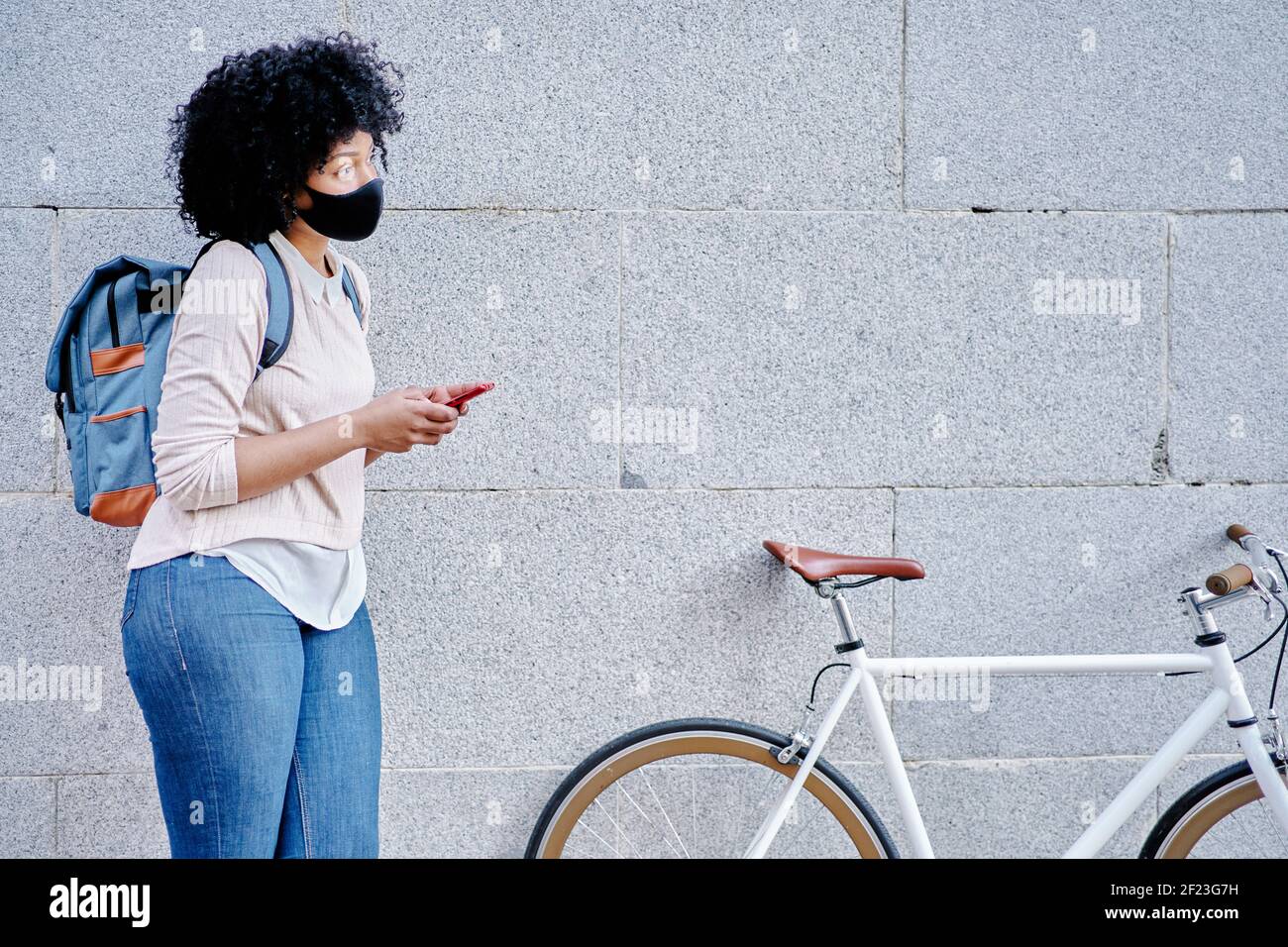 Donna afroamericana con uno smartphone in mano. Donna con maschera protettiva. Andare in bicicletta in città. Foto di alta qualità Foto Stock
