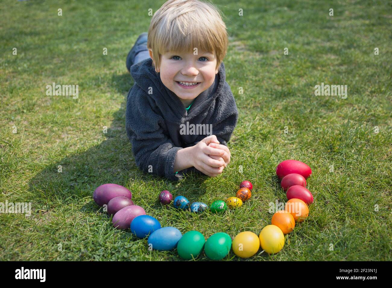 Il bambino felice giace sull'erba vicino al pollo e alle uova di quaglia, coloratissimi in colori arcobaleno, soleggiato giorno di primavera. Tradizioni familiari, gioioso bambino Foto Stock