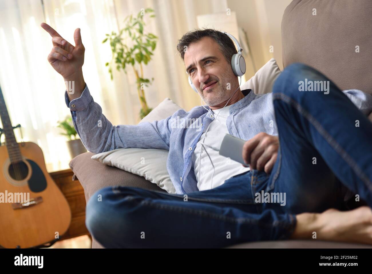 Felice uomo adulto che danzava in divano ascoltando musica da un cellulare con cuffie bianche vestite casual soggiorno seguendo il ritmo o Foto Stock