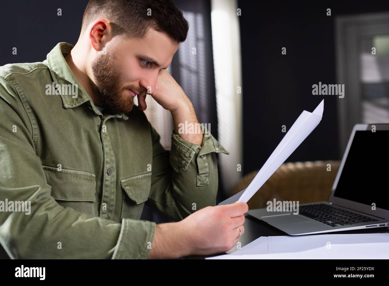 Uomo caucasico che guarda stanco guardando i documenti mentre si siede al tavolo del computer. Documenti, lavoro e lavoro online. Foto Stock