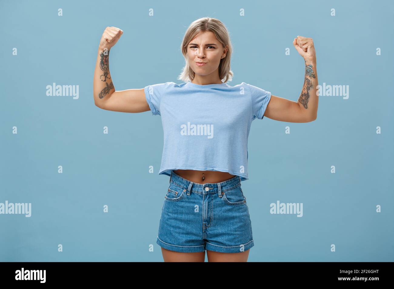 Donne sportive forti e potenti con tatuaggi in t-shirt e. shorts alzando le braccia mostrando grandi muscoli e bicipiti puzzano Foto Stock