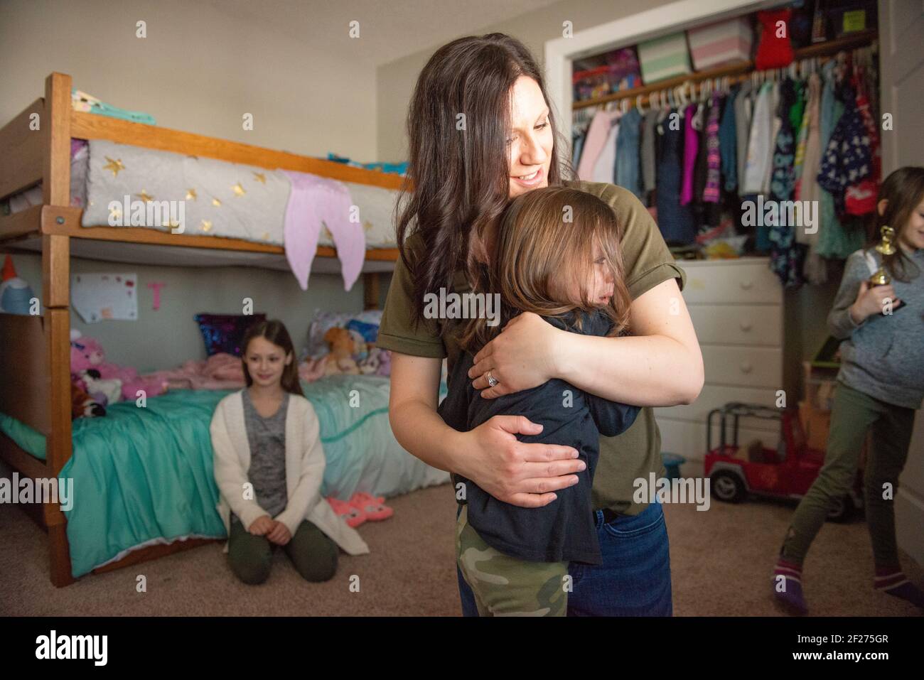 Mamma che abbraccia un bambino piangente mentre le sorelle guardano sopra. Foto Stock