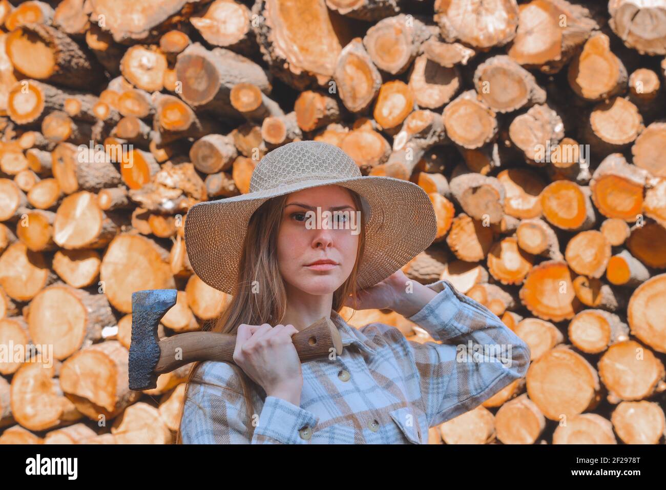 Ritratto esterno di giovane bella donna con capelli lunghi, ascia in mano, con cappello di paglia, in elegante camicia lumberjack su sfondo di legno, persone. Foto Stock