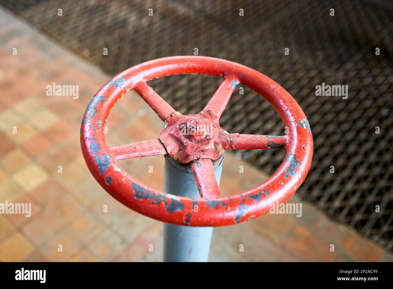 Vecchia ruota della valvola rossa verniciata arrugginita con rigonfiamenti e spellature messa a fuoco selettiva con dispositivo di estensione sullo sfondo fuori fuoco pavimento piastrellato e. Foto Stock