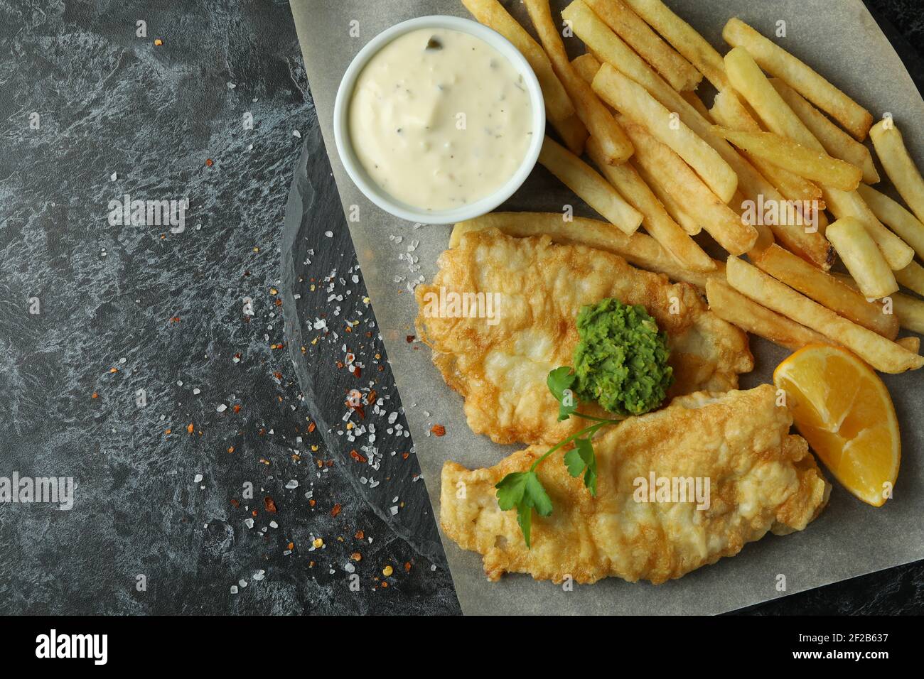 Carta da forno con pesce fritto e patatine su un tavolo nero fumoso, pasto rustico da asporto Foto Stock