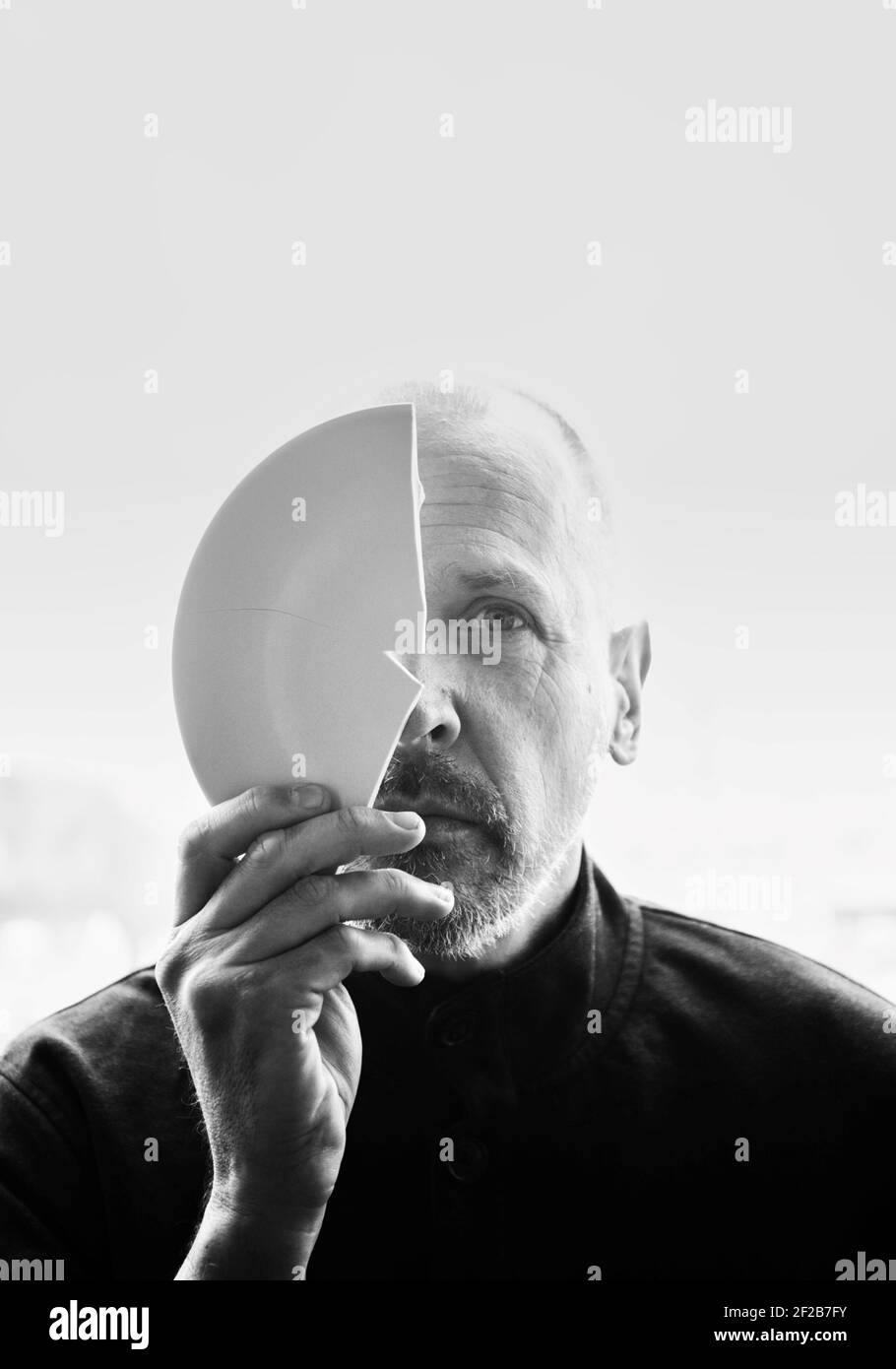 Immagine in bianco e nero di un uomo medio adulto che tiene un piatto rotto che copre metà del viso. Concetto di mistero, intrigo, curiosità Foto Stock