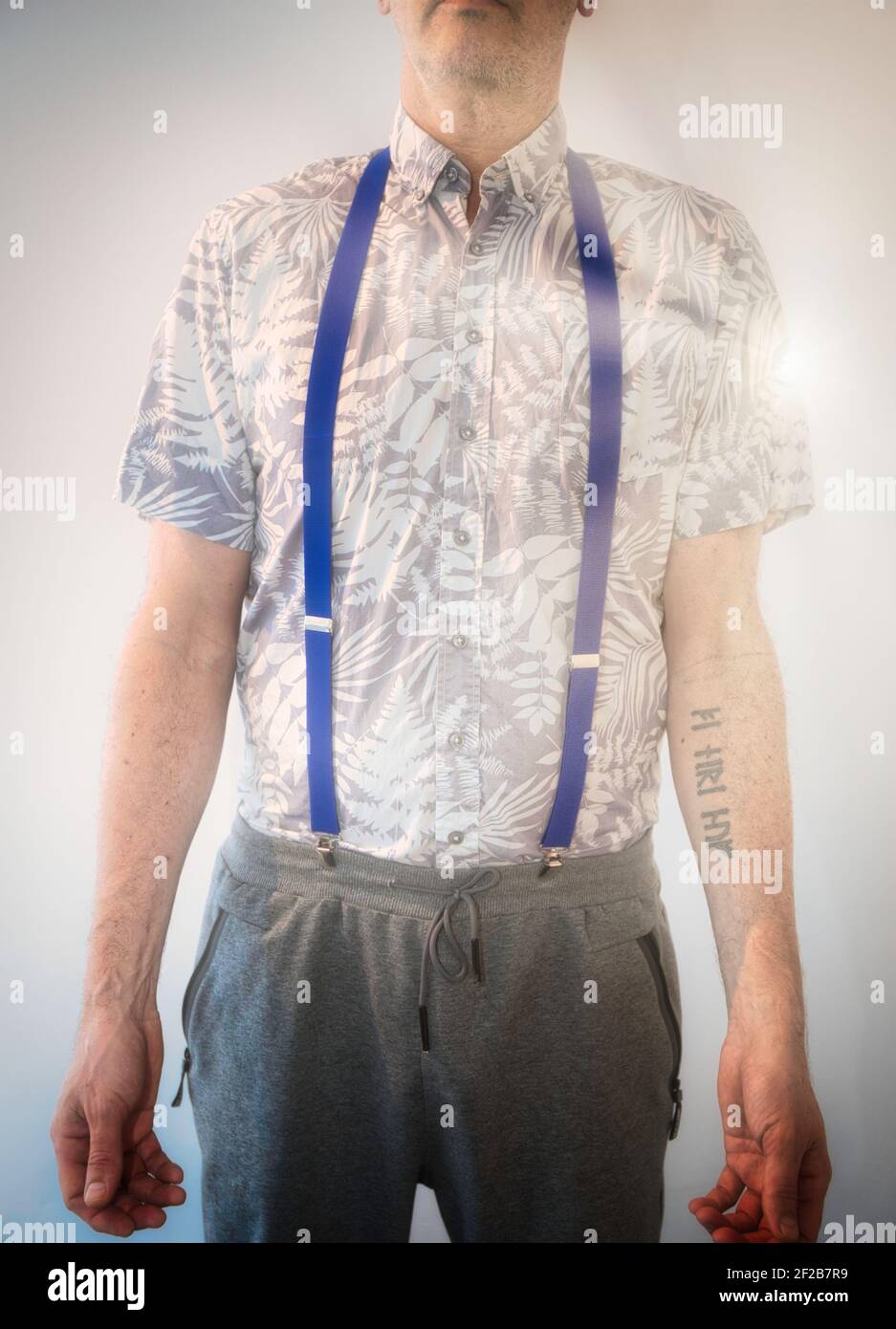 Uomo medio adulto con tatuaggio con braccio in piedi alla luce del sole che indossa sospenditori, camicia estiva e pantaloni sudore. Concetto di gente, moda, stile di vita Foto Stock