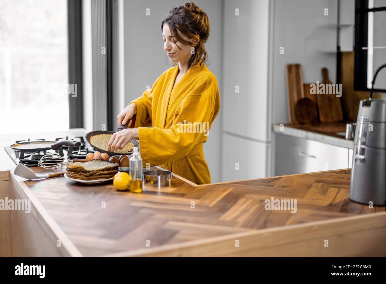 Donna felice vestita in accappatoio giallo cuoce frittelle per la colazione, mentre si trova vicino al piano cottura nella moderna cucina interna a casa Foto Stock